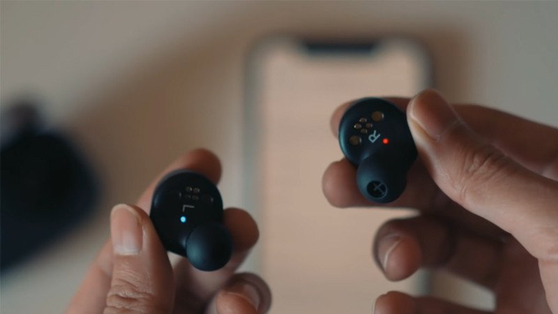 Bàn tay nhấn giữ nút nguồn của tai nghe Bluetooth màu đen, với đèn báo đang nhấp nháy, thể hiện bước khởi động tai nghe để kết nối với điện thoại Samsung.